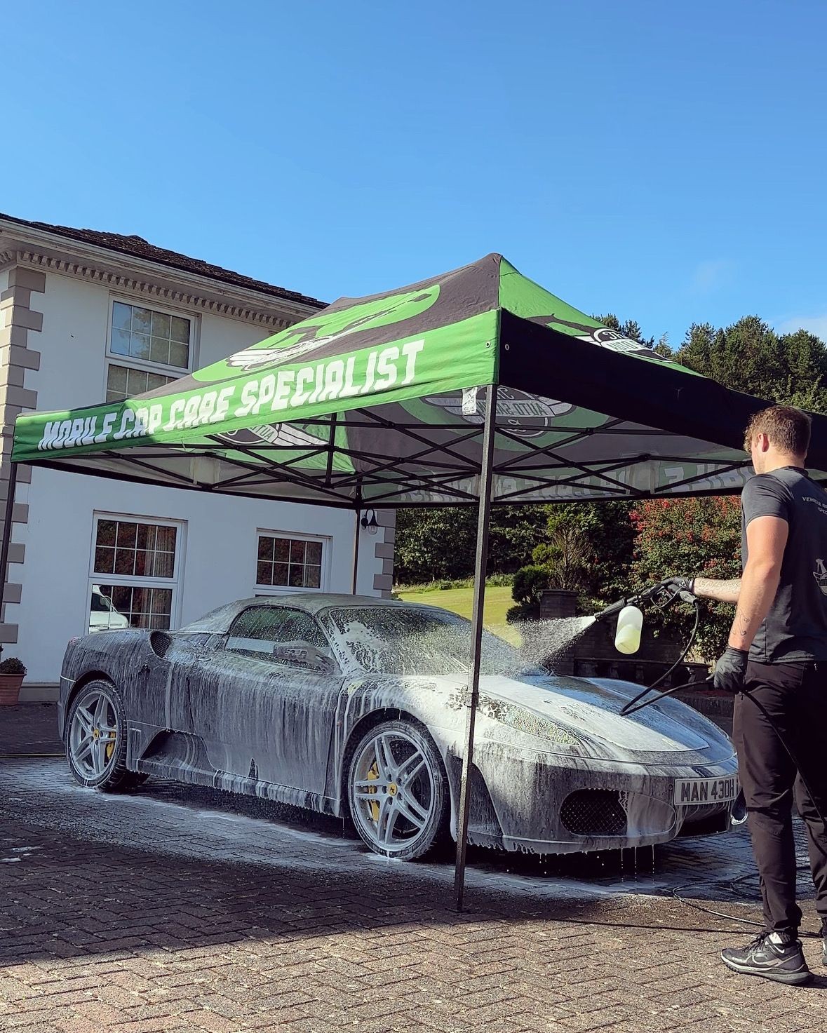Person washing a sports car covered in foam under a car care specialist tent outside a house.