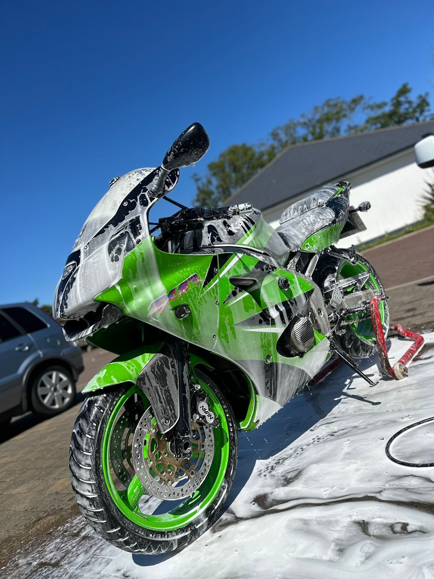 Green motorcycle covered in soap foam during a wash, parked outdoors on a sunny day.