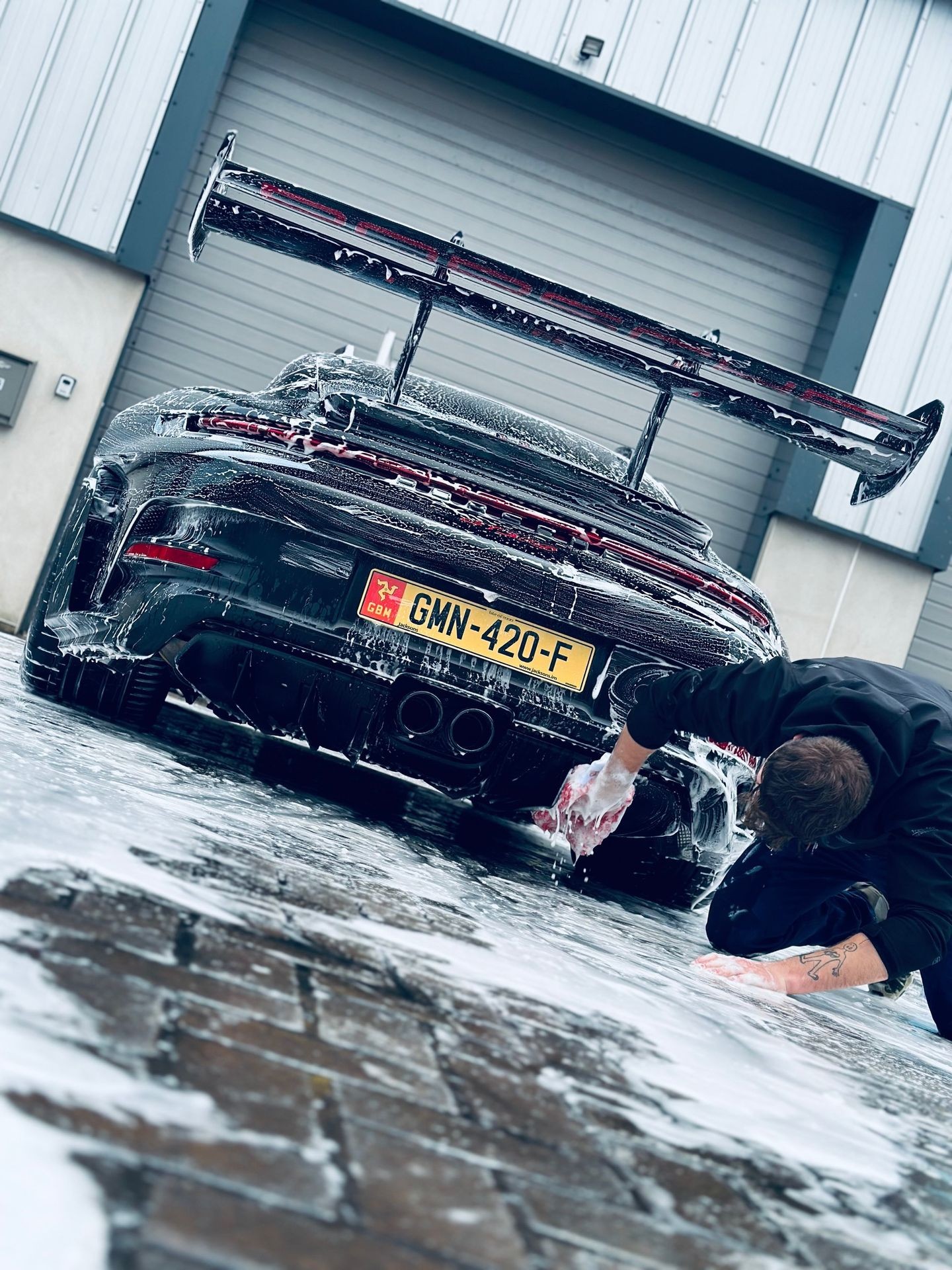 Person washing the rear of a sports car covered in soap suds outside a garage.