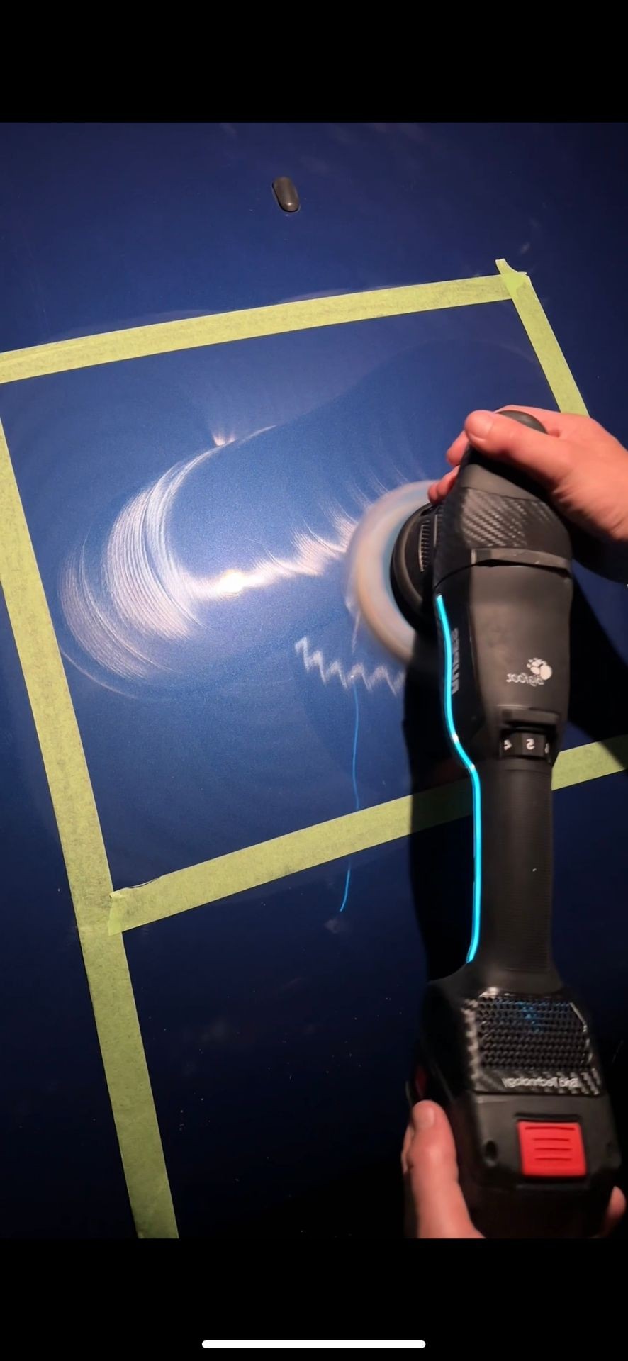 Person using a car polisher on a blue painted surface with green tape borders.