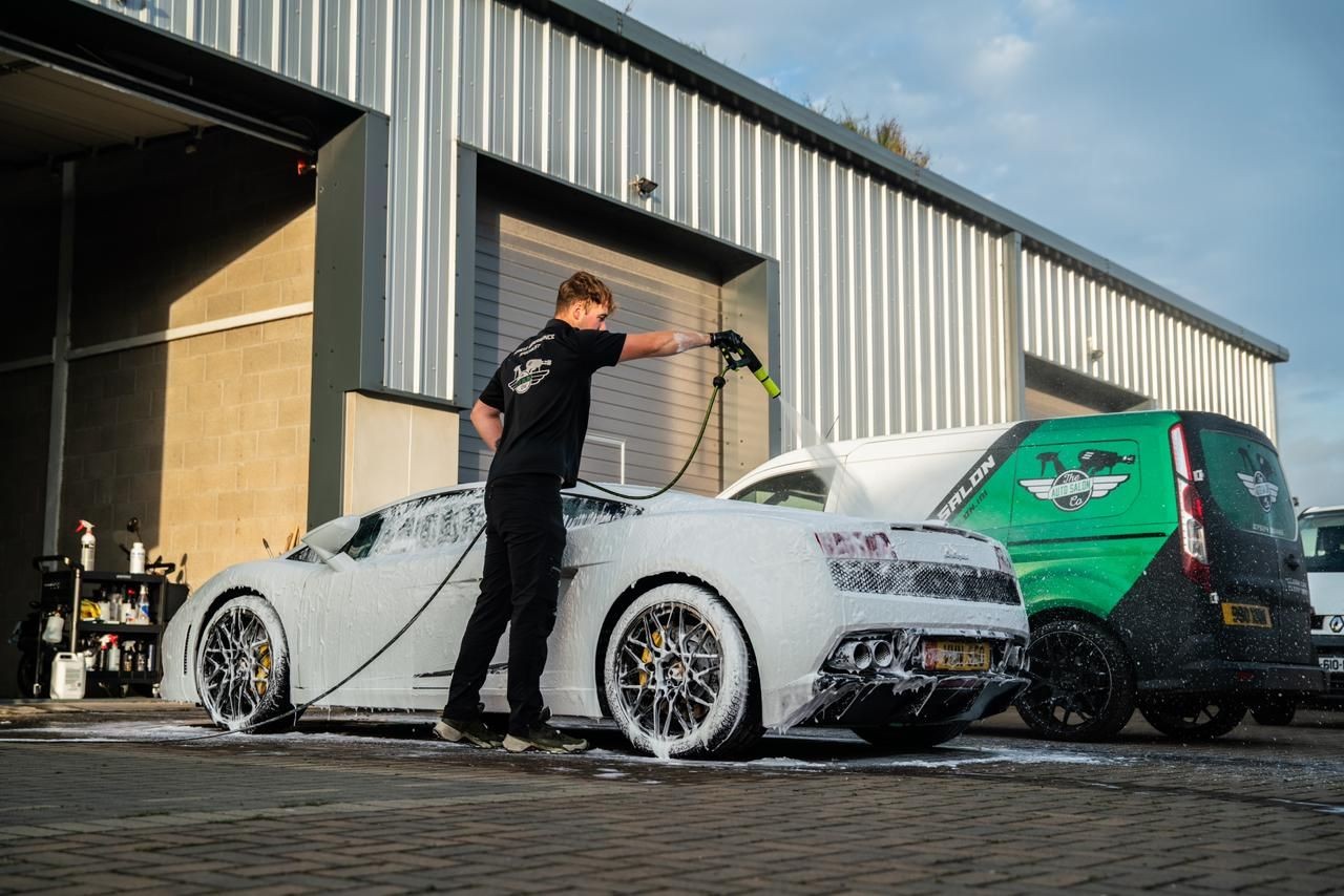 Person washing sports car with foam outside a garage, next to a green van.