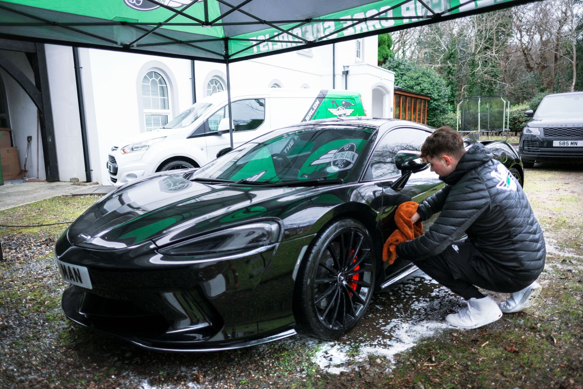 Person cleaning a sleek black sports car under a canopy, surrounded by other vehicles and building structures.