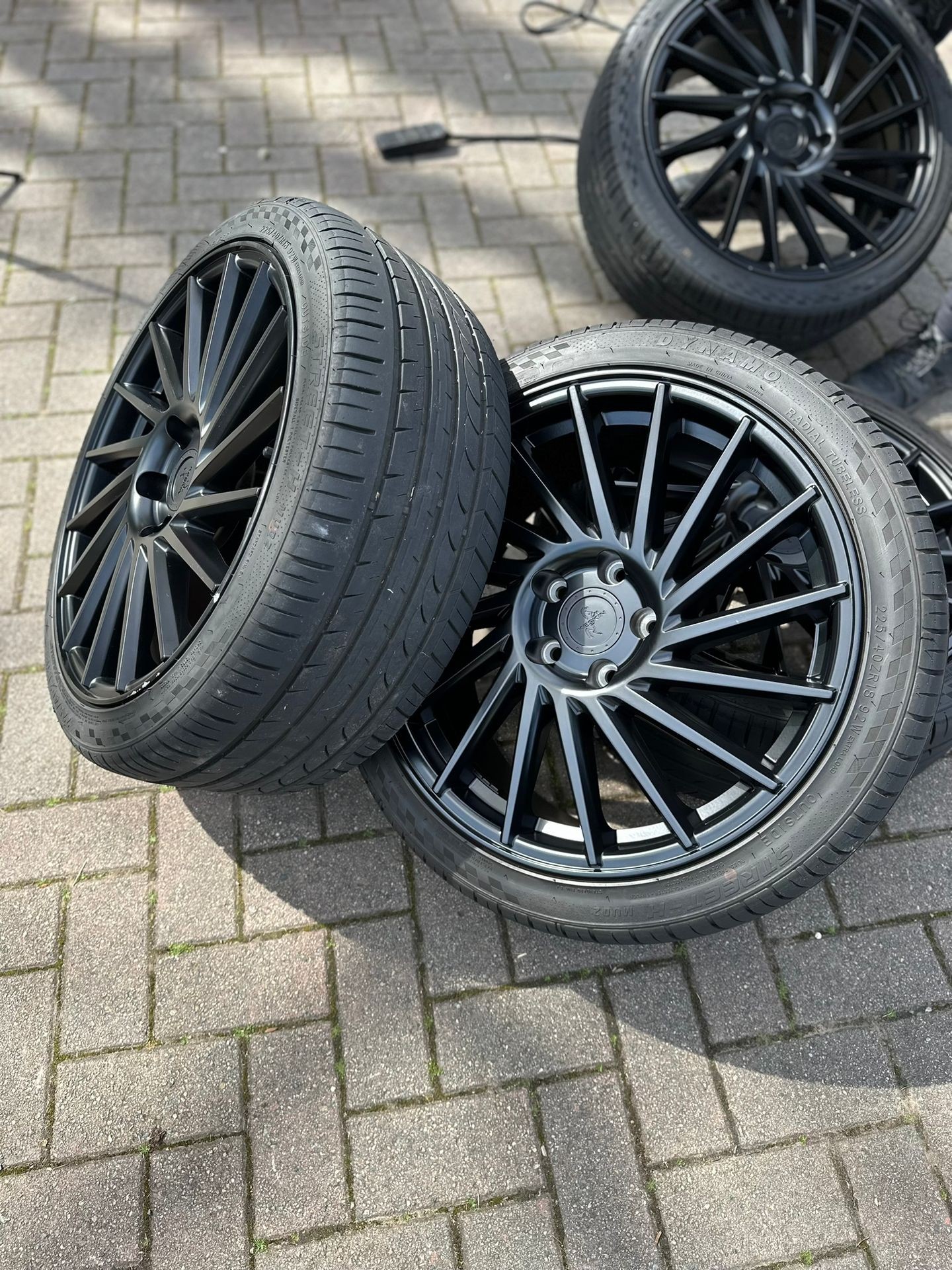 Close-up of two black alloy car wheels with tires, positioned on a brick pavement.
