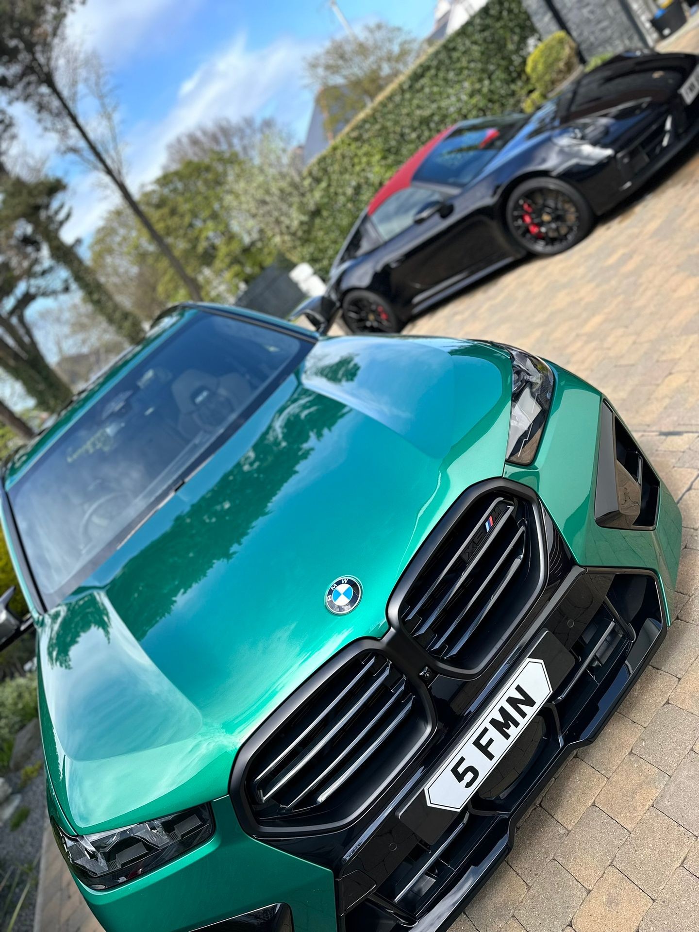 Green BMW and black Porsche parked on a paved driveway with trees and hedges in the background.